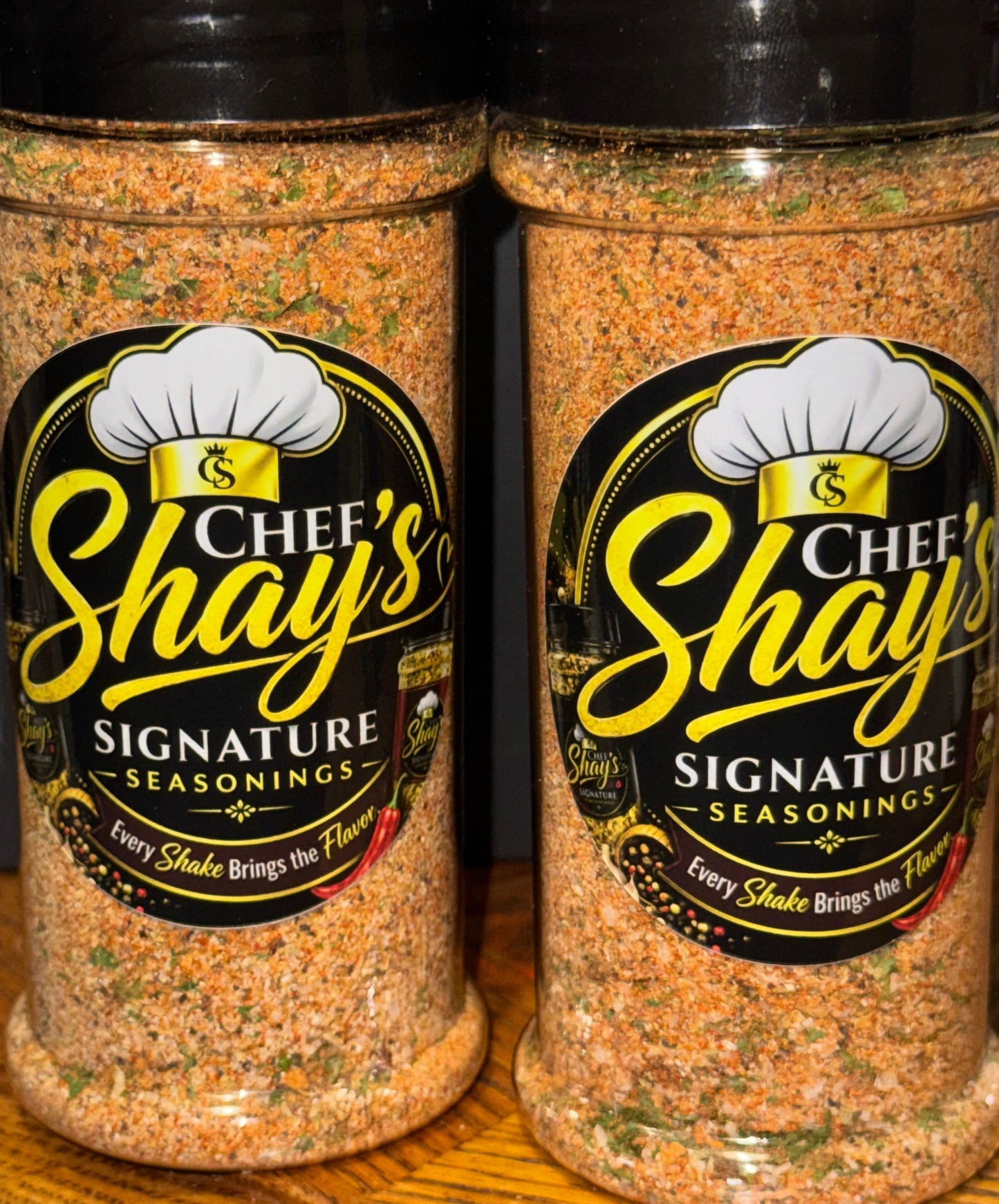 Two clear plastic bottles of Chef's Shay Signature Seasonings filled with a spice blend, resting on a wooden surface