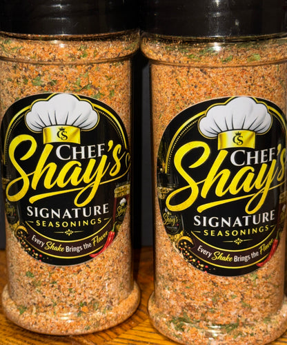Two clear plastic bottles of Chef's Shay Signature Seasonings filled with a spice blend, resting on a wooden surface
